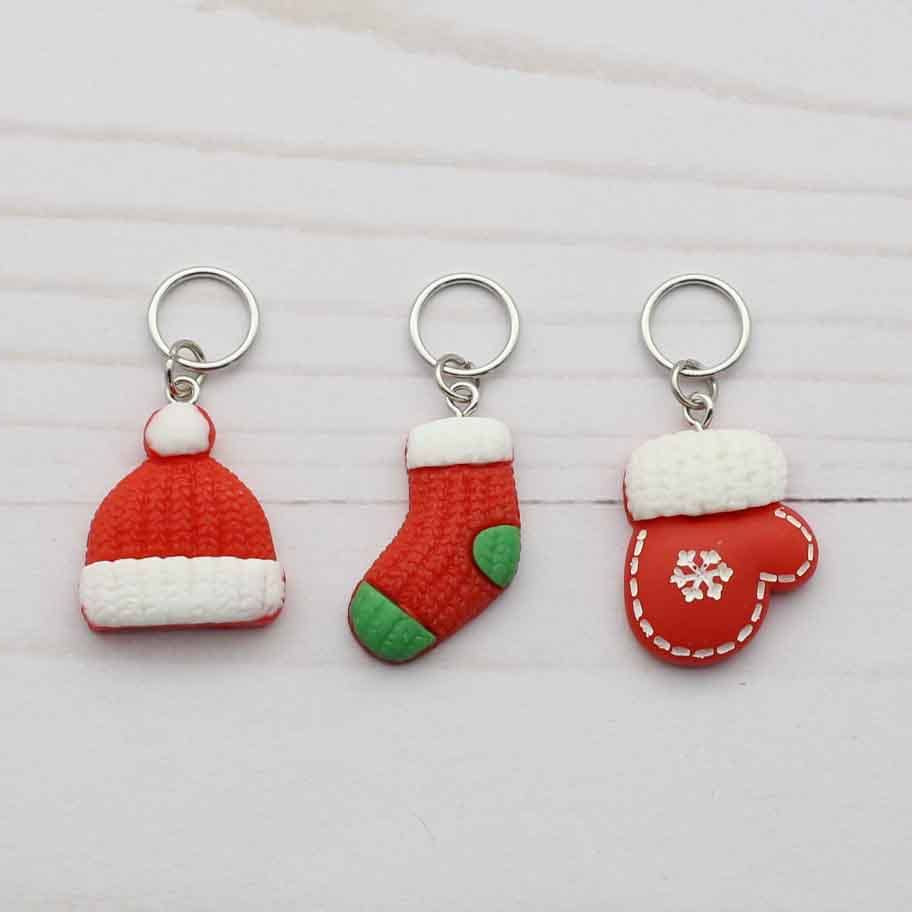 Three small Christmas-themed stitch markers on a light wood background: a red and white Santa hat, a red and green stocking, and a red mitten with a white snowflake.